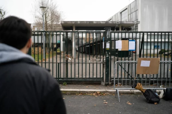 Grilles fermées devant un lycée bloqué, avec des barrières de fortune et quelques effets d’élèves au premier plan.
