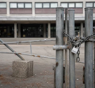 Grilles fermées d’un lycée public bloqué, chaînes et barrières improvisées devant un portail sobre, façade en arrière-plan.