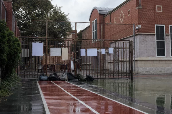 Entrée bloquée d’un lycée derrière des grilles fermées, sous la pluie, avec façade scolaire et sacs d’élèves au sol.