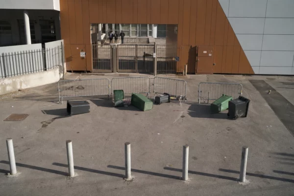 Entrée de lycée bloquée par des poubelles renversées et des barrières, sous une lumière d’hiver rasante.