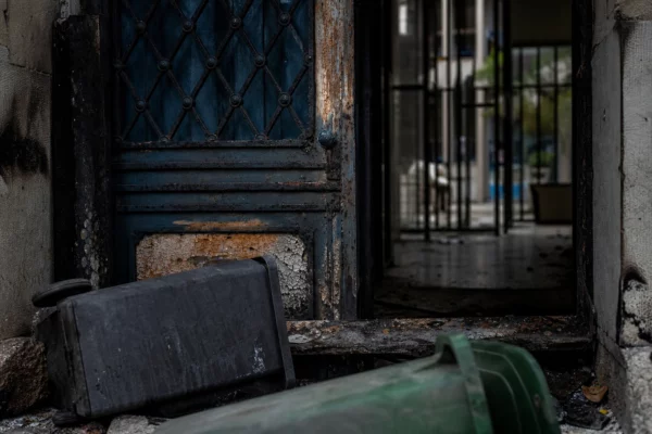 Porte de lycée noircie par un incendie, poubelles renversées au premier plan, façade scolaire en arrière-plan.