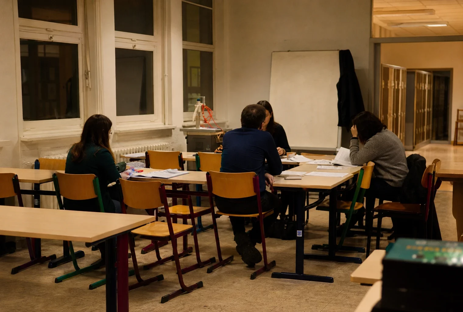 Salle de lycée française vide, tables et chaises regroupées au premier plan, tableau et fenêtres au fond, lumière chaude du soir.