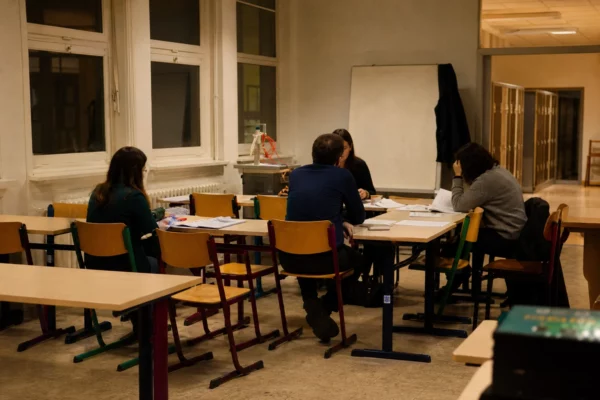 Salle de lycée française vide, tables et chaises regroupées au premier plan, tableau et fenêtres au fond, lumière chaude du soir.