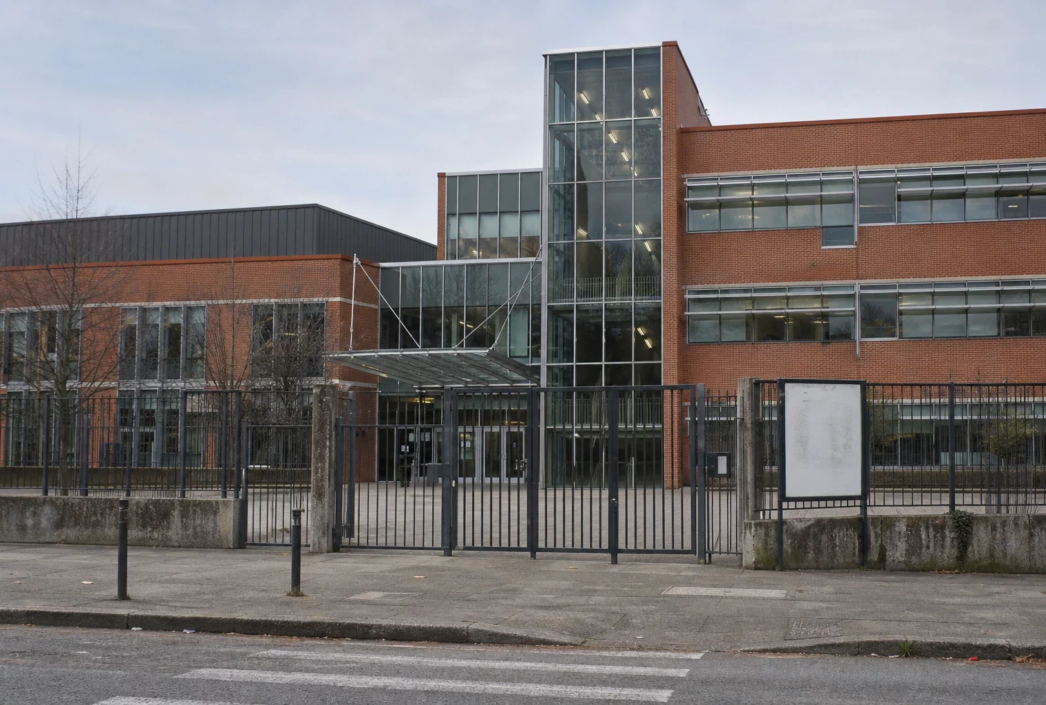 Entrée d’un lycée public français avec grilles, portail et façade en brique et verre, sous une lumière douce.