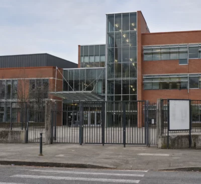 Entrée d’un lycée public français avec grilles, portail et façade en brique et verre, sous une lumière douce.