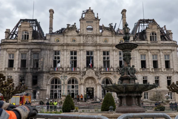 Vue de l’hôtel de ville de Limoges après l’incendie, avec au premier plan des mains gantées posées sur une barrière de sécurité de chantier, devant les vestiges calcinés de la mairie et les débris de toiture.