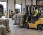 Des palettes de matériel militaire protégé et sanglé sont déplacées par un chariot dans un entrepôt logistique en activité.