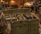 Caisses militaires ouvertes contenant du matériel de soutien sanglé dans une zone de stockage, avec rayonnages et autres caisses en arrière-plan.