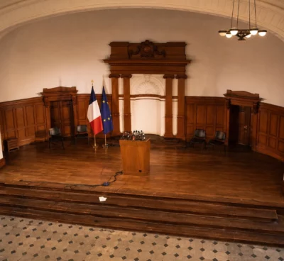 Salle officielle de Matignon vide, pupitre central et micros, quelques chaises au fond, ambiance de conférence sous lumière chaude.