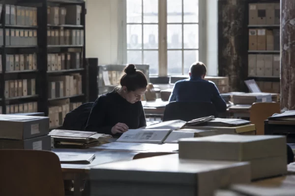 Des notices d’enquêtes électorales imprimées, avec des tableaux de paramètres, sont consultées sur une table près de boîtes d’archives.
