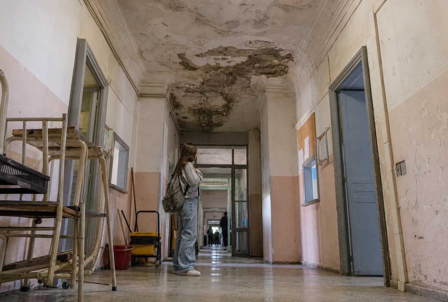 Couloir de lycée public avec plafond qui fuit, traces d’humidité, portes de classes et matériel scolaire abandonné.