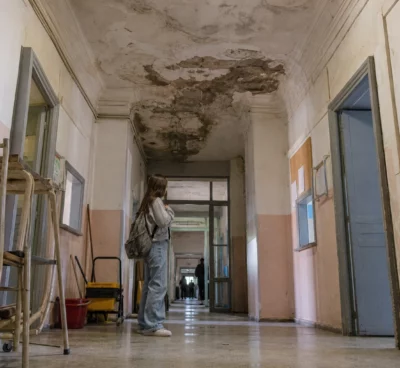 Couloir de lycée public avec plafond qui fuit, traces d’humidité, portes de classes et matériel scolaire abandonné.