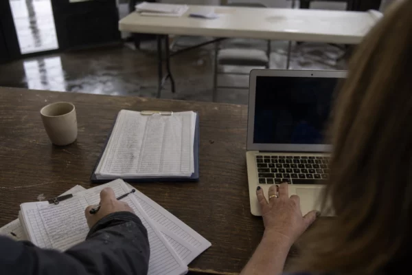 Bureau de permanence avec ordinateur ouvert, main sur le clavier et feuilles d’inscription, sous une lumière grise et pluvieuse.