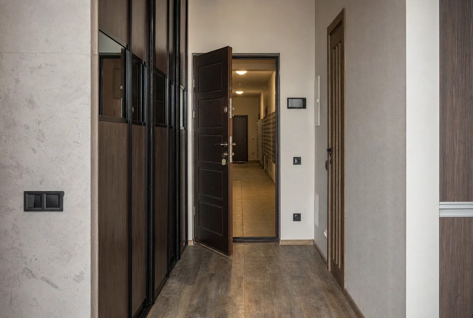 Porte d’appartement entrouverte dans un hall résidentiel, couloir vide avec boîtes aux lettres et traces d’usage.