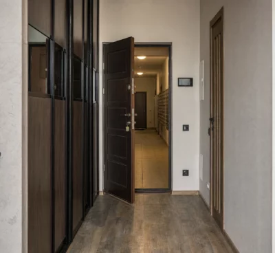 Porte d’appartement entrouverte dans un hall résidentiel, couloir vide avec boîtes aux lettres et traces d’usage.