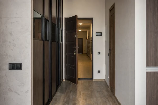 Porte d’appartement entrouverte dans un hall résidentiel, couloir vide avec boîtes aux lettres et traces d’usage.