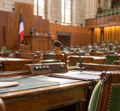 Gros plan sur des pupitres en bois usés, équipés de microphones et de boîtiers de vote, dans un hémicycle parlementaire.
