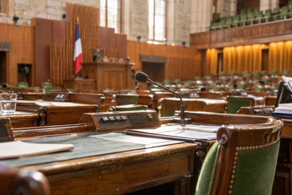 Gros plan sur des pupitres en bois usés, équipés de microphones et de boîtiers de vote, dans un hémicycle parlementaire.