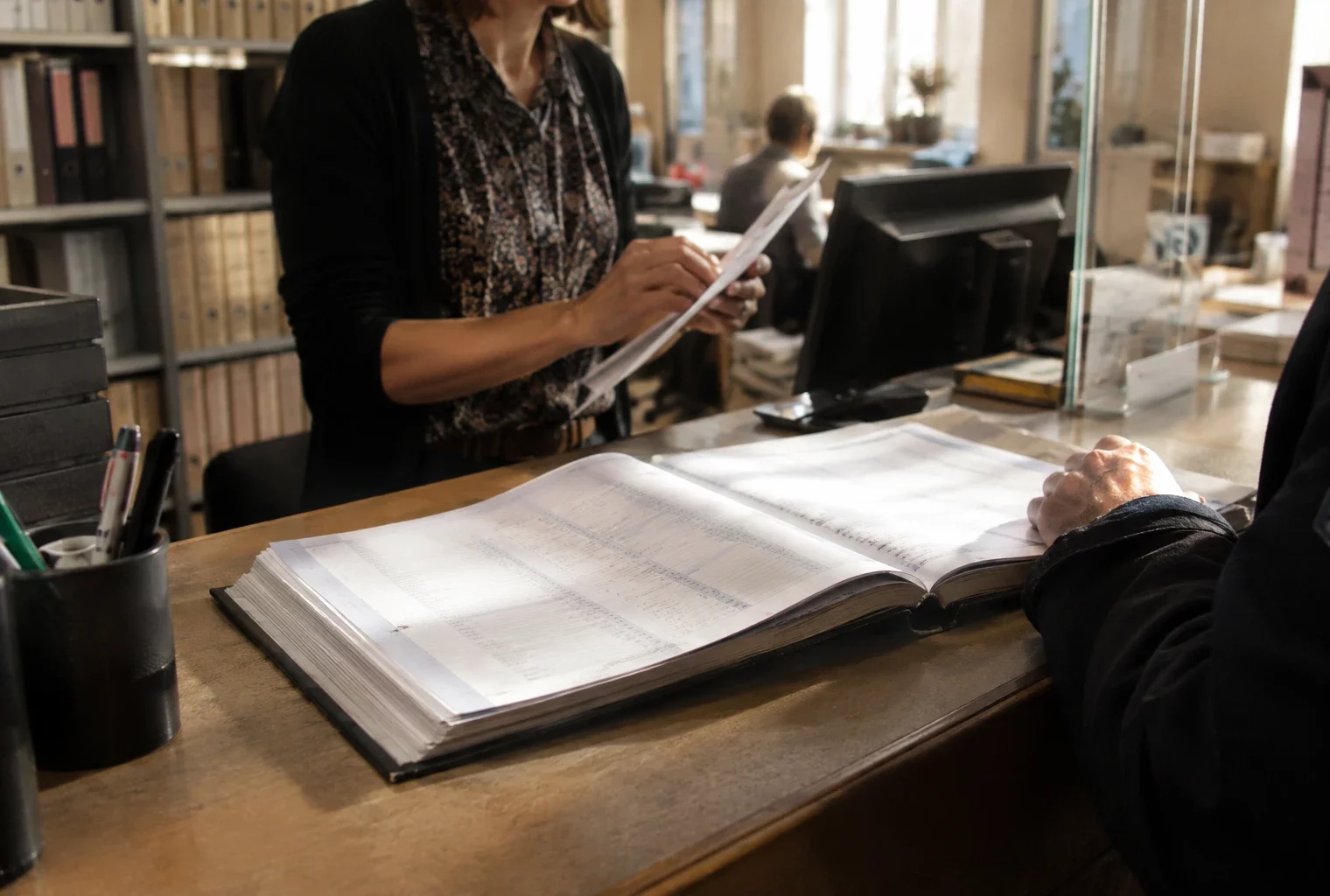 Registre électoral ouvert sur le comptoir d’un service municipal, parmi des livres et dossiers administratifs aux pages indistinctes.