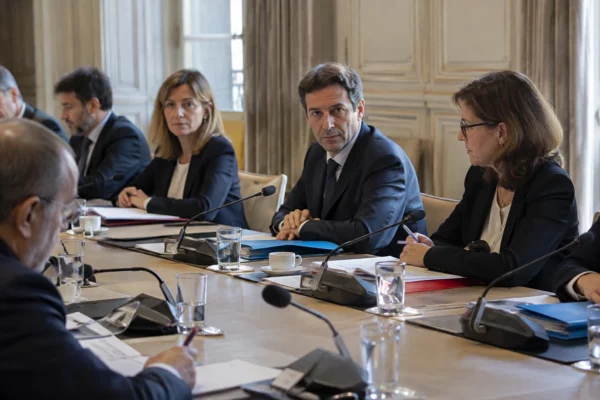Salle de réunion institutionnelle avec une longue table, des microphones de table, des dossiers sobres et plusieurs sièges.