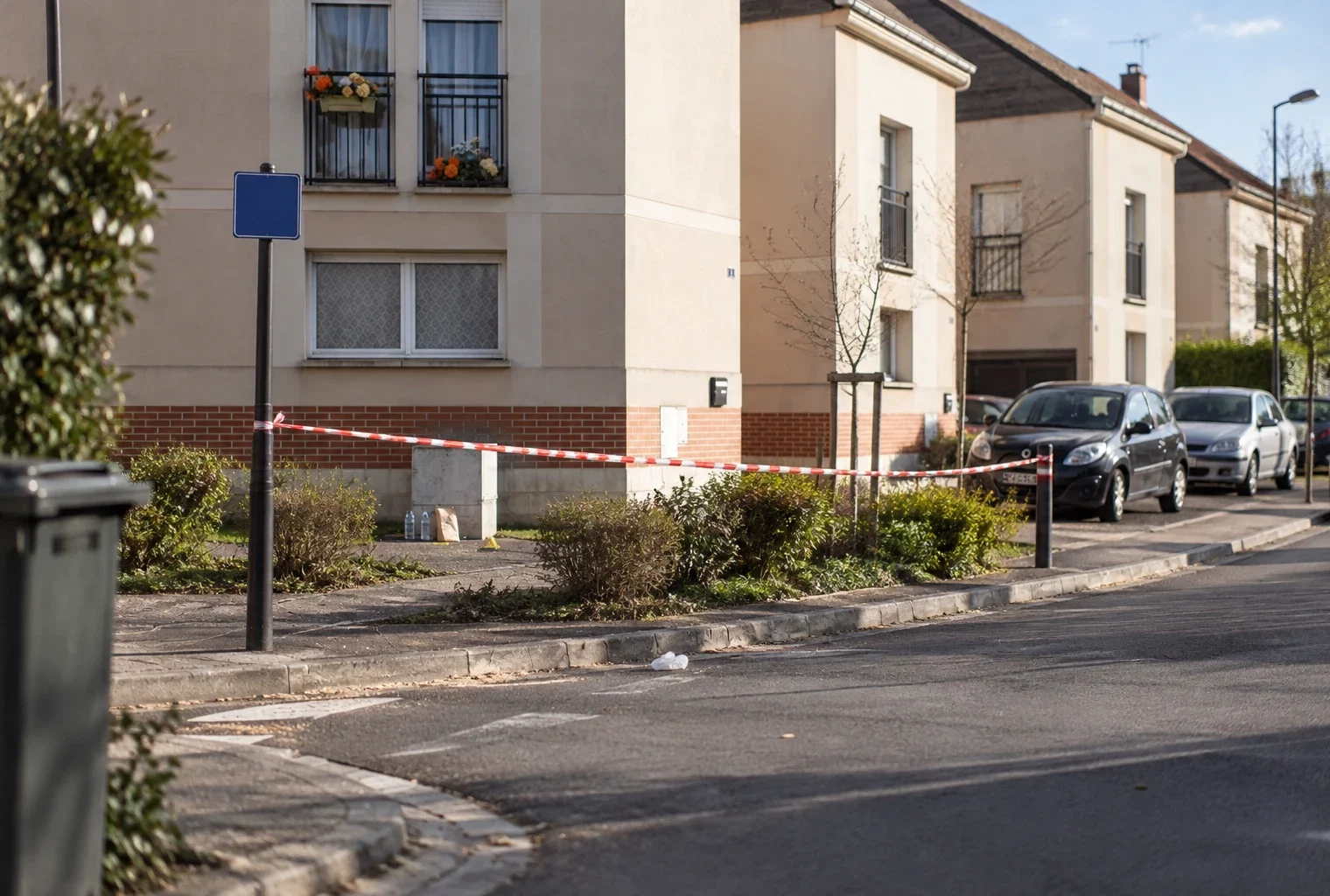 Une rue résidentielle ensoleillée, avec un ruban de sécurité discret et des traces d’intervention près du trottoir.