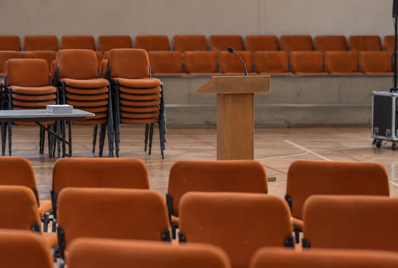 Salle polyvalente vide avec chaises empilables, un pupitre équipé d’un microphone et une rangée de sièges au fond.