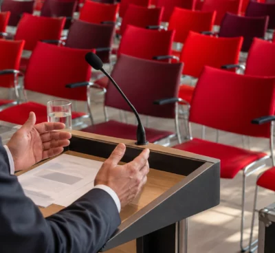 Des mains posées sur un pupitre avec microphone, devant des rangées de chaises rouges dans une salle de conférence.