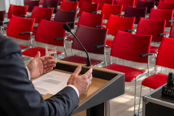 Des mains posées sur un pupitre avec microphone, devant des rangées de chaises rouges dans une salle de conférence.