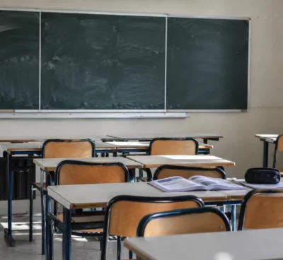 Salle de classe vide avec des tables d’élèves, des chaises et un tableau mural, sans inscription lisible ni personne.