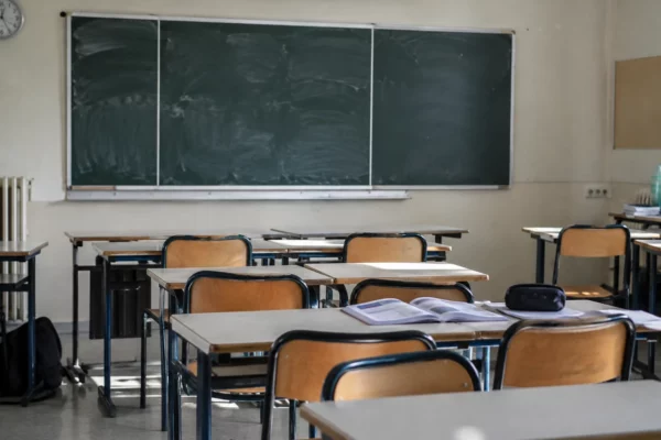 Salle de classe vide avec des tables d’élèves, des chaises et un tableau mural, sans inscription lisible ni personne.