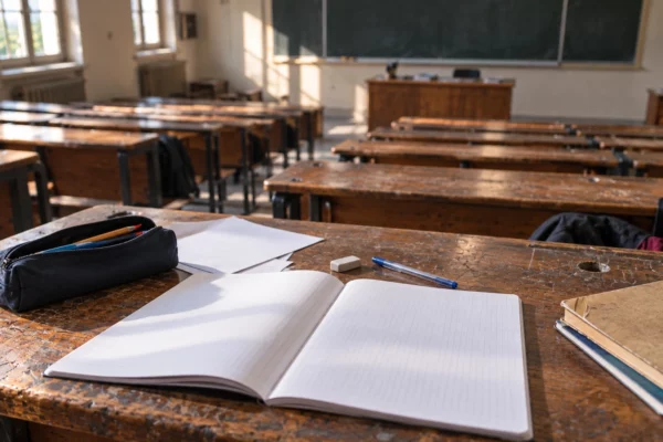 Gros plan sur une table d’élève usée, avec cahier vierge et fournitures, dans une salle de classe aux rangées de tables.