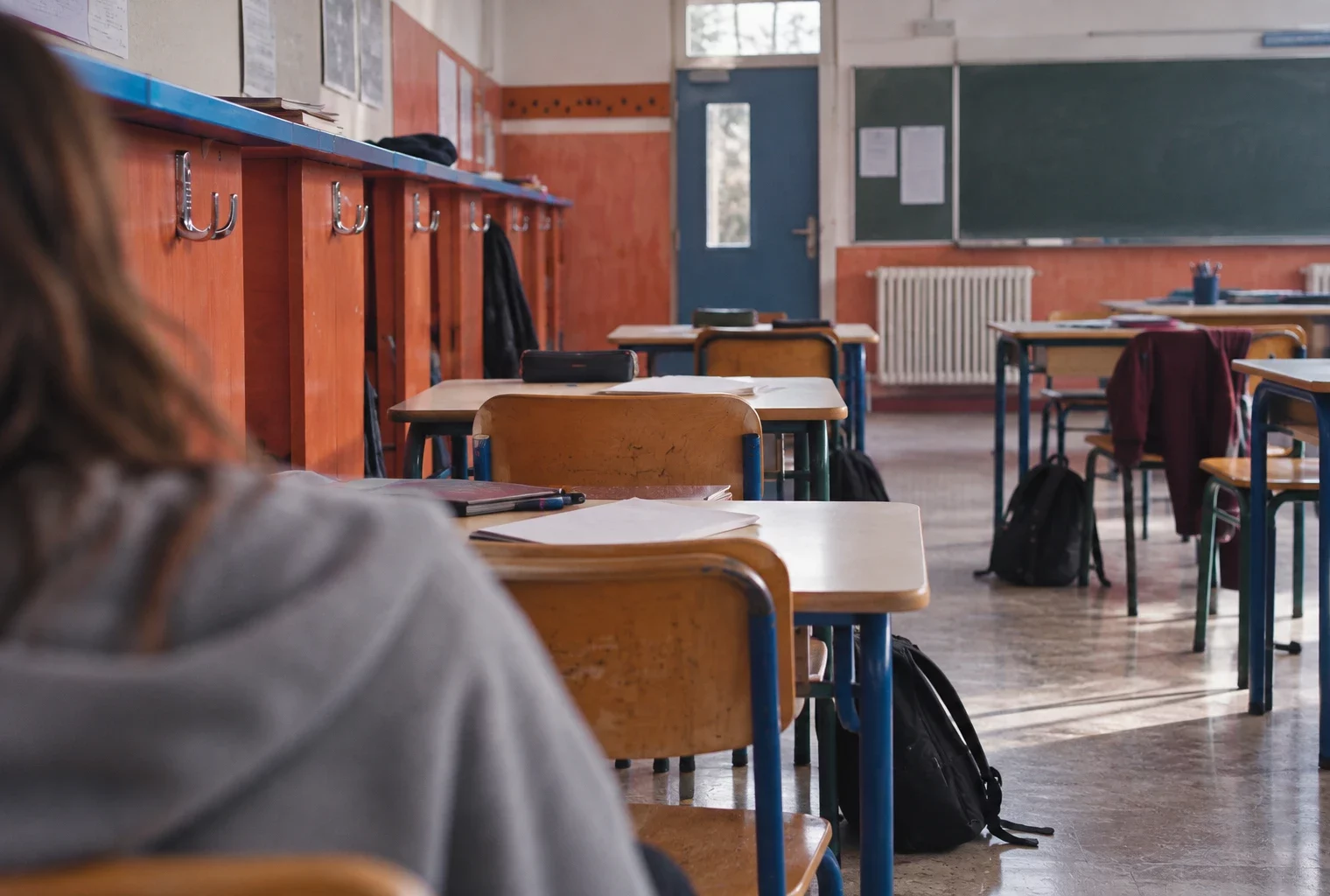 Salle de classe de lycée avec tables usées, chaises dépareillées, cahiers laissés sur les bureaux et casiers en bois.