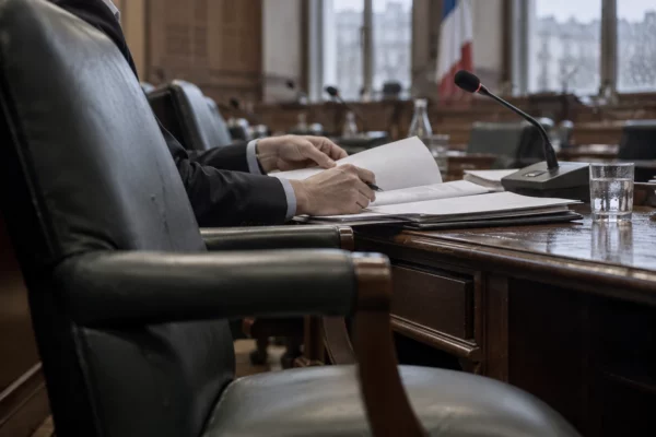 Dans une salle de commission, des mains consultent des dossiers vierges près d’un microphone, devant des rangées de pupitres en bois.