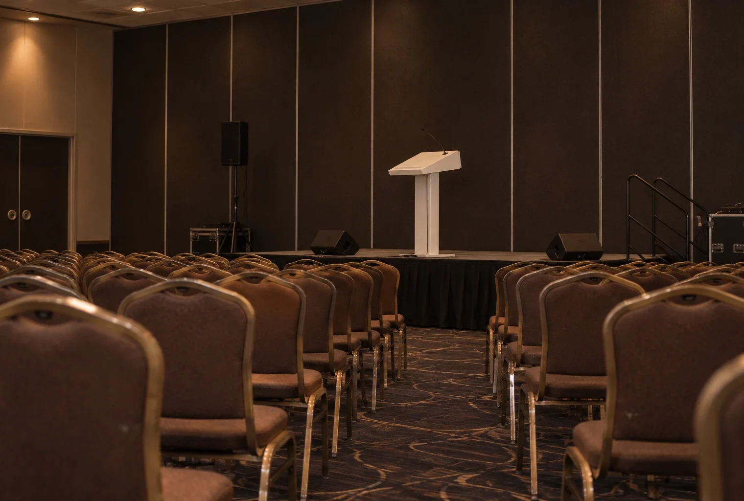 Salle de congrès vide, avec rangées de chaises usées face à une estrade et un pupitre équipé d’un microphone.