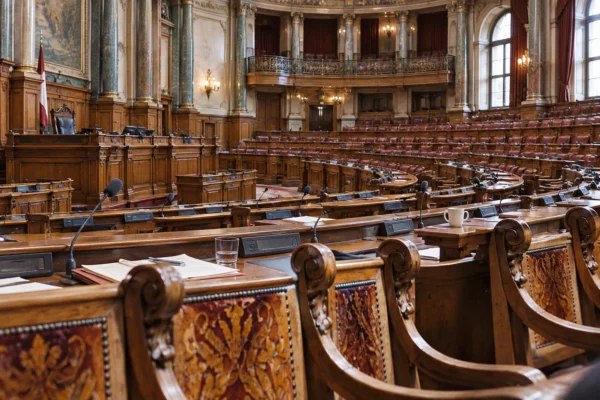Rangées de pupitres équipés de microphones au milieu de sièges en bois sculpté dans une salle parlementaire.