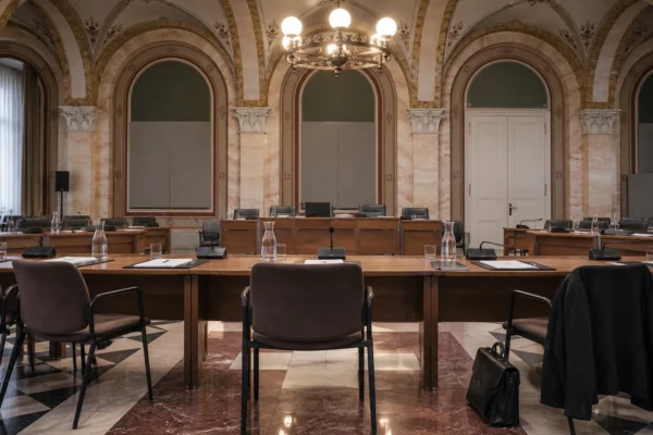 Salle de conseil municipale aux arches de pierre, avec pupitres, microphones et dossiers vierges devant des rangées de sièges.