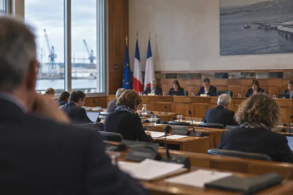 Vue en profondeur d’une salle de conseil municipal, avec pupitres en bois, microphones et sièges de conseillers.