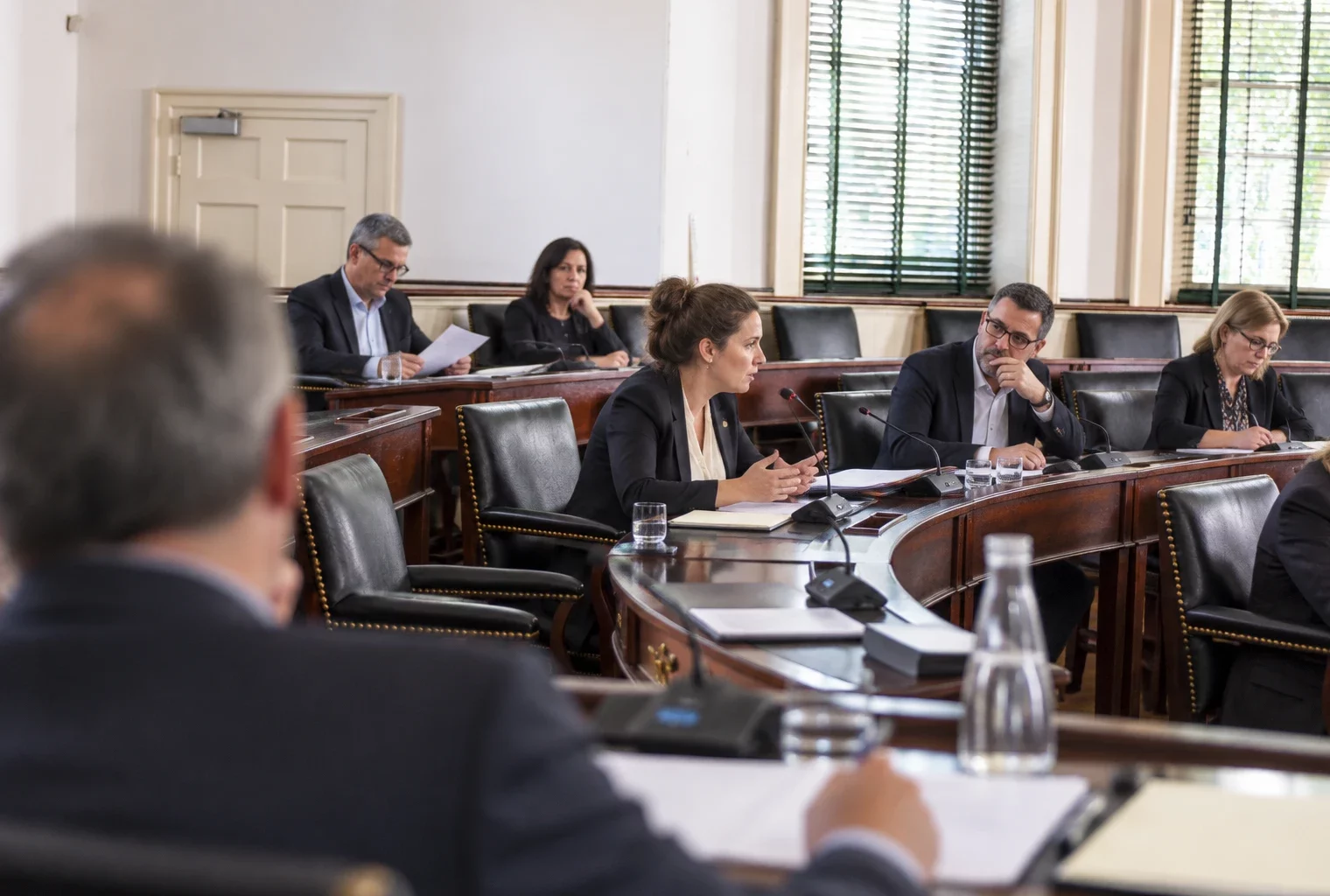 Dans une salle de conseil aux bureaux de bois usés, des élus délibèrent autour de dossiers et de microphones.