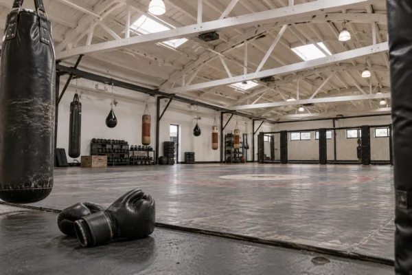 Salle d’entraînement de MMA vide, avec tapis usé, gants posés au sol et sacs de frappe sous une charpente apparente.