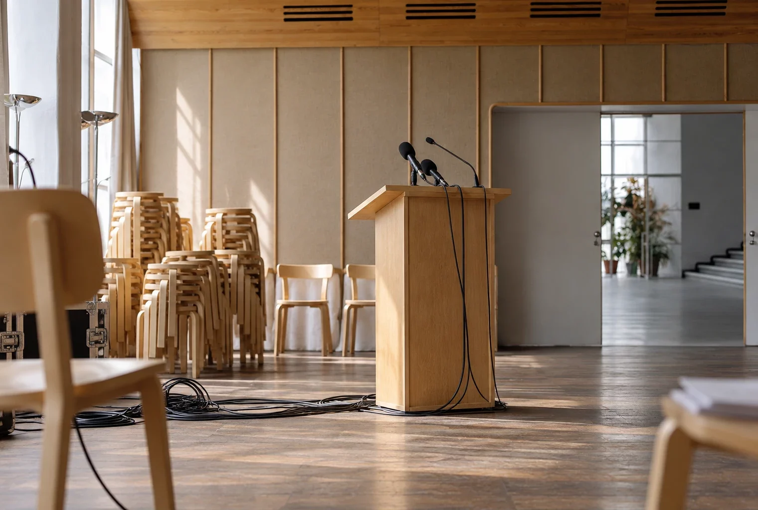 Chaises empilées et pupitre équipé de microphones et de câbles dans une salle municipale aux murs acoustiques et au sol usé.