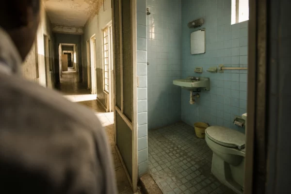 Sanitaires vétustes d’un lycée public, carrelage abîmé, lavabo usé et couloir scolaire en arrière-plan.