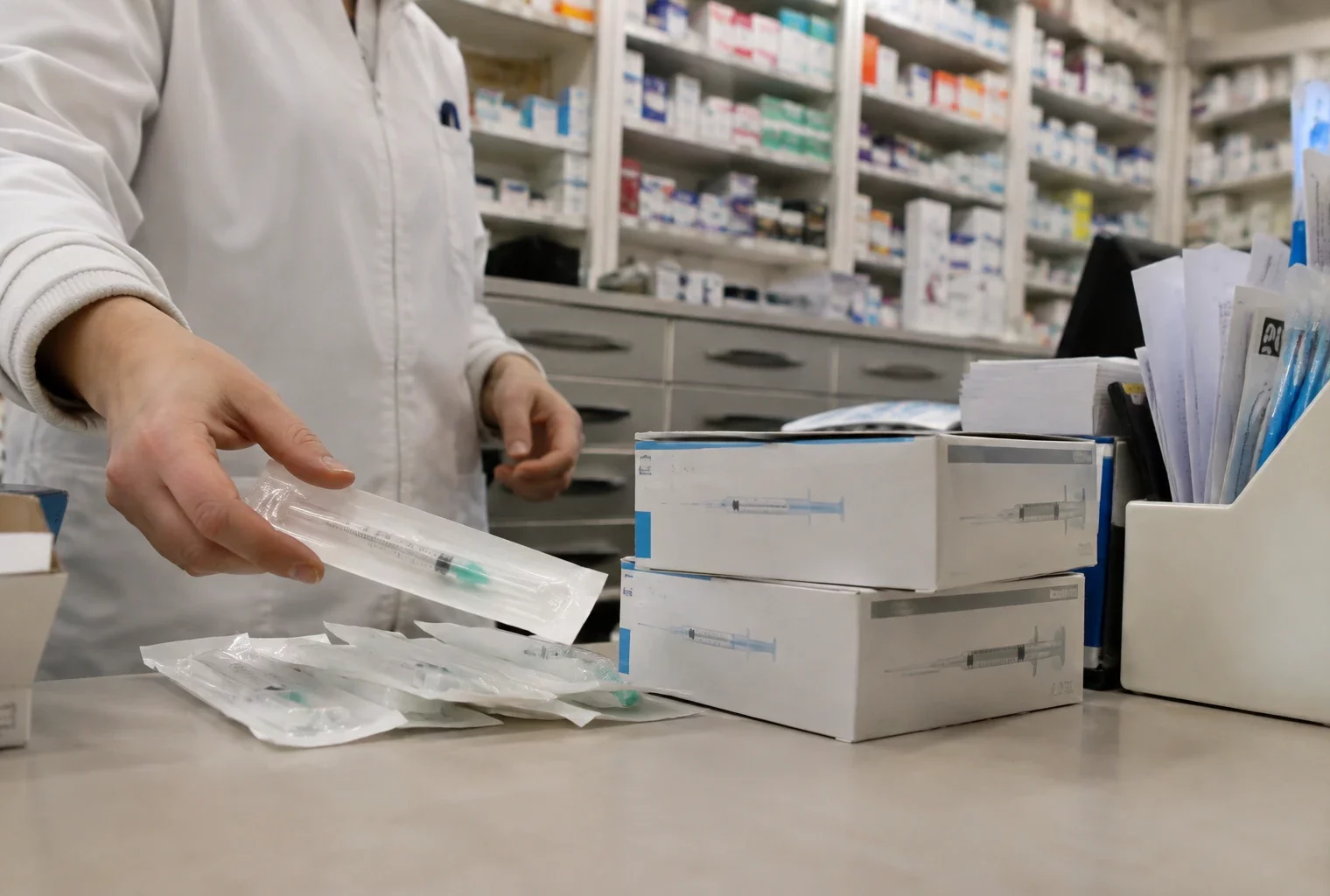 Boîtes de seringues médicales scellées sur un comptoir de pharmacie, avec des tiroirs de rangement visibles en arrière-plan.