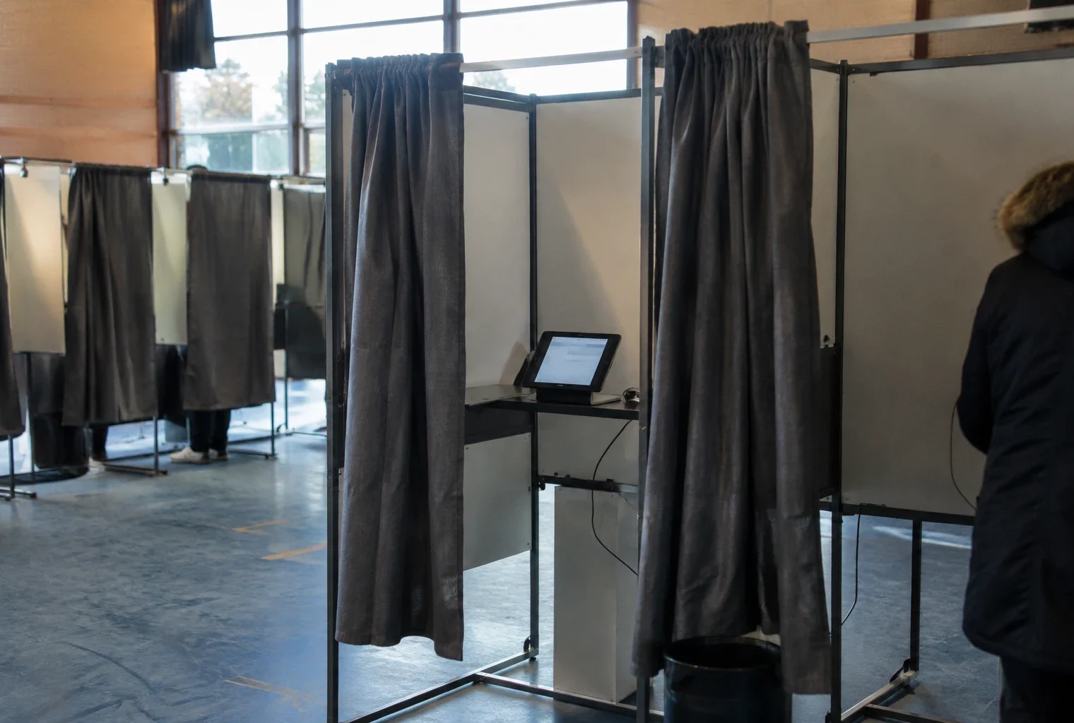 Une tablette est installée dans un isoloir, au milieu de parois de vote et d’autres cabines dans une salle polyvalente.