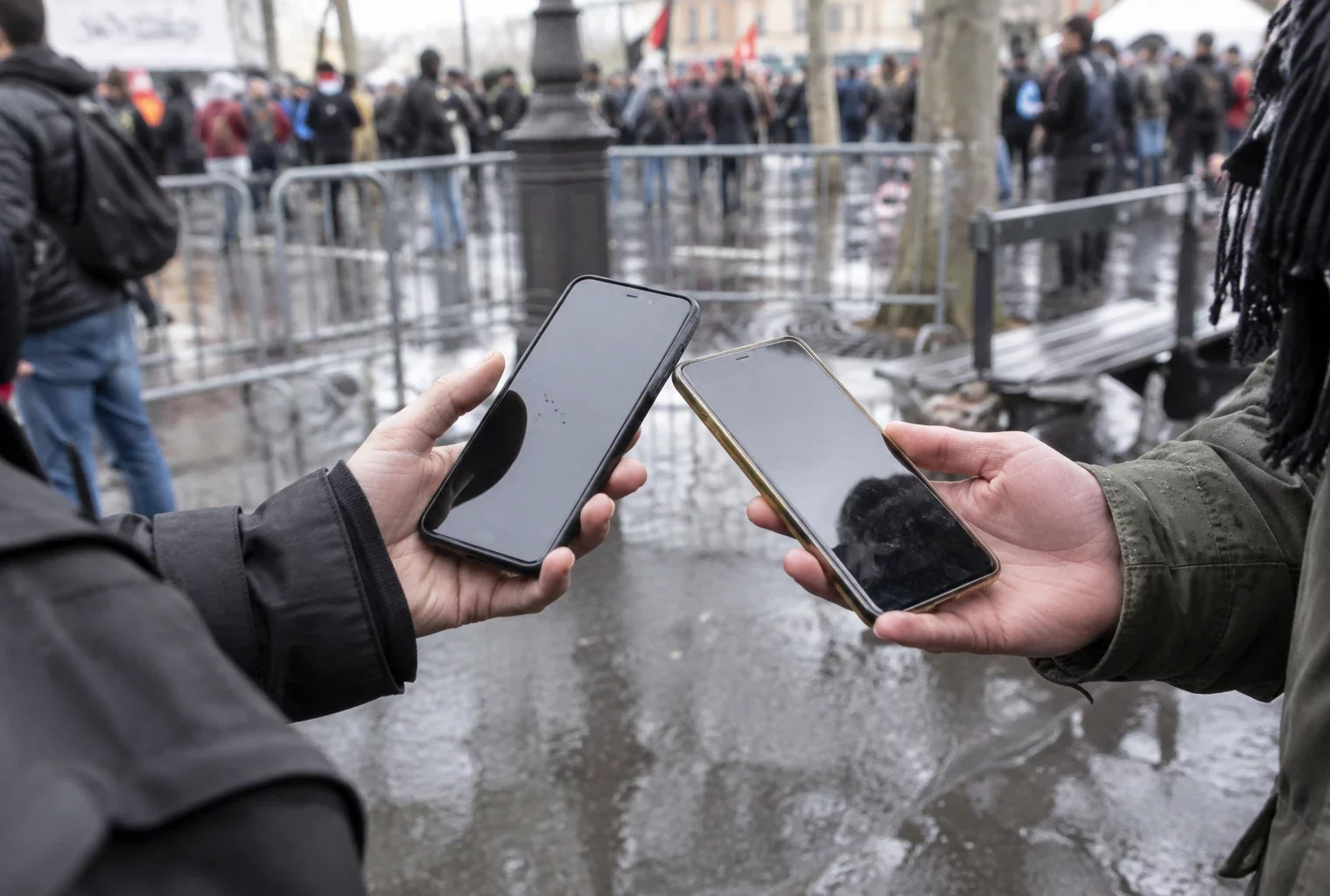 Deux personnes rapprochent leurs téléphones sur un trottoir, devant des barrières métalliques et des silhouettes floues.