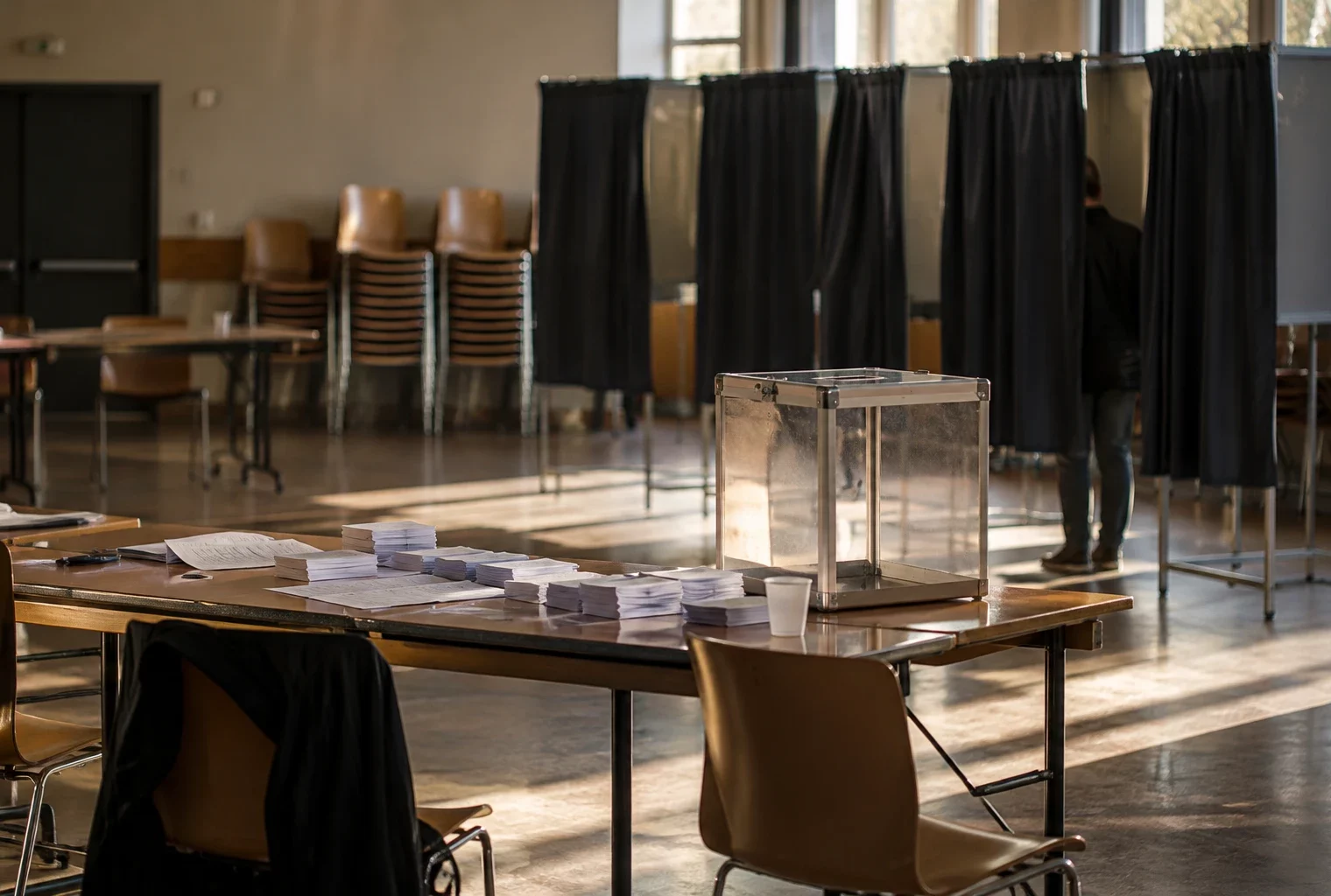 Urne fermée et bulletins empilés sur une table de dépouillement, avec des isoloirs et des chaises rangées en arrière-plan.