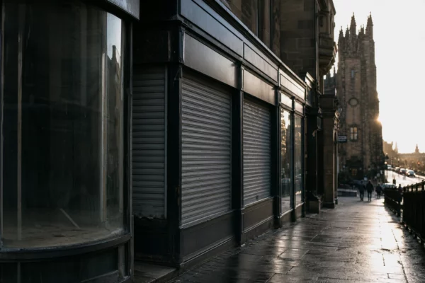 Façades de boutiques fermées, rideaux baissés et vitrines vides dans une rue commerçante française, en lumière dure de contre-jour.