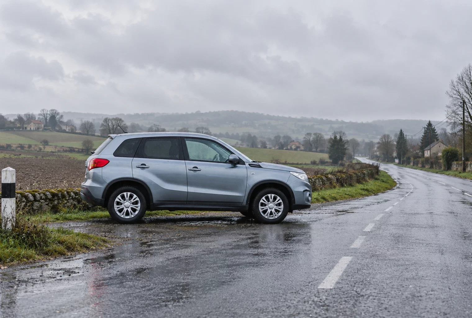 Une voiture ordinaire stationnée au bord d’une route rurale mouillée, entre champs et maisons espacées sous un ciel gris.