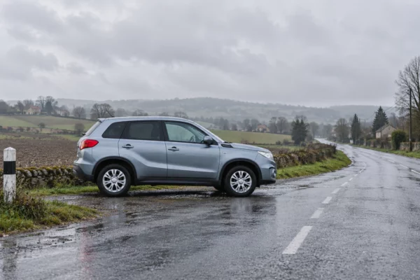 Une voiture ordinaire stationnée au bord d’une route rurale mouillée, entre champs et maisons espacées sous un ciel gris.