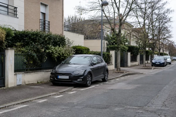 Une voiture est stationnée au bord d’une rue résidentielle, devant des façades d’immeubles et du mobilier urbain.