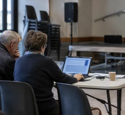 Un ordinateur portable affiche une interface de vote neutre dans une salle municipale avec des chaises empilées en arrière-plan.
